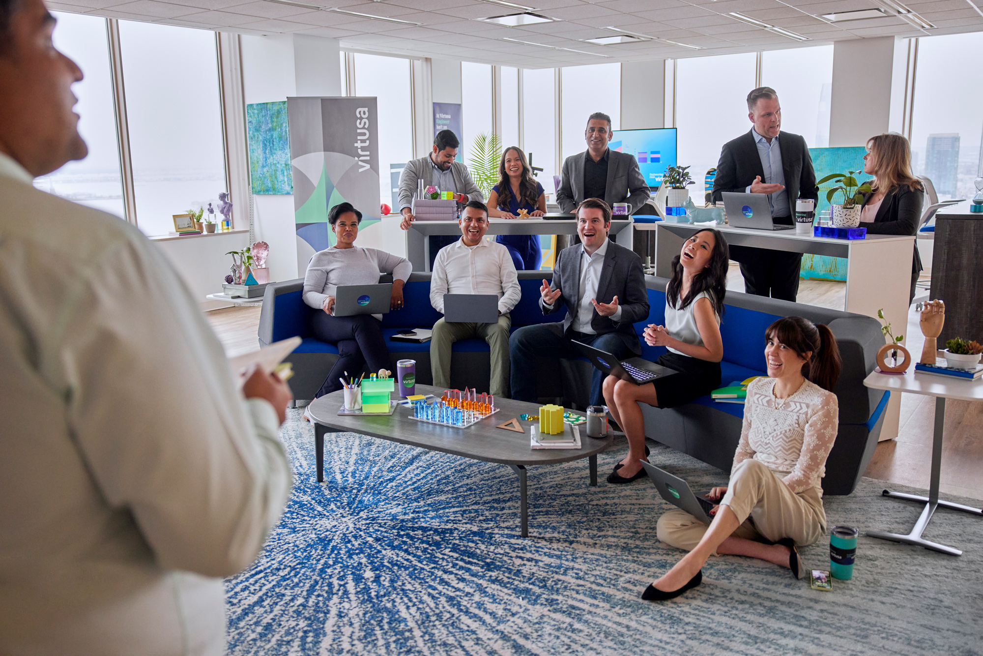 Office meeting with diverse group around tables, some using laptops, and stationary supplies on a table.